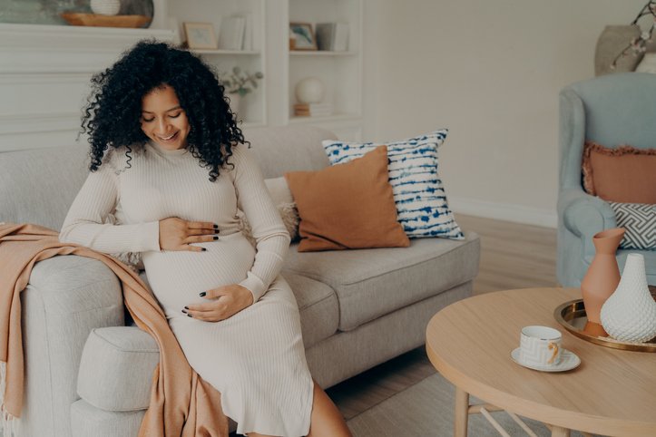 Smiling beautiful pregnant mixed race woman sitting on couch and touching her belly